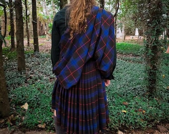May include: A person wearing a traditional Scottish kilt and plaid. The kilt is a dark blue and black tartan with red lines, paired with a matching plaid draped over the shoulder. The person is wearing black boots and gray socks.