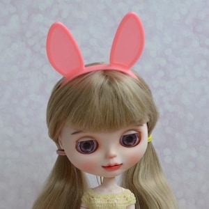 Può includere: Una bambola con lunghi capelli biondi, che indossa una fascia con orecchie da coniglio rosa e un vestito giallo all'uncinetto. La bambola ha grandi occhi viola e guarda avanti.