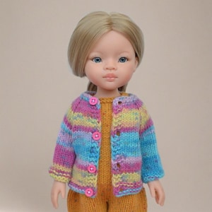 May include: A doll wearing a colorful, striped knit cardigan with pink buttons. The cardigan features shades of blue, purple, and yellow. The doll is also wearing a mustard-colored outfit. The doll has blonde hair and blue eyes.