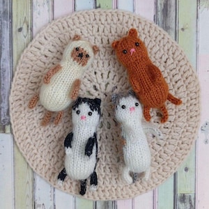 Può includere: Quattro figurine di gatti lavorati a maglia a mano in vari colori: bianco con dettagli marroni, arancione, bianco e nero e bianco con dettagli grigi. I gatti sono su un tappeto circolare all'uncinetto color crema.