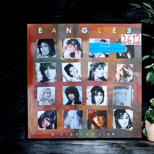 Puede incluir: Portada del álbum "Different Light" de The Bangles. La portada presenta un collage de fotos cuadradas de los miembros de la banda, con el nombre de la banda en la parte superior y el título del álbum en la parte inferior. Es visible una pegatina con el texto "Manic Monday".