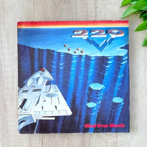 Peut inclure: Pochette d'album vinyle vintage du groupe 220 Volt. La pochette présente une illustration futuriste avec un vaisseau spatial, des personnages et le texte "Mind Over Muscle". La palette de couleurs comprend des nuances de bleu, de rouge et de blanc.