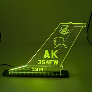 May include: A neon green acrylic light shaped like an aircraft tail fin. The fin features the letters "AK" and "354FW", along with a state outline and a crest. The light sits on a black base and is illuminated with a bright green glow.