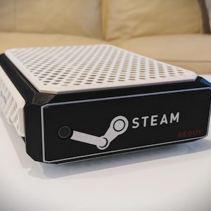 Peut inclure: Console Steam Redux noire et blanche avec un dessus perforé blanc. Le logo Steam et le mot « STEAM » en blanc sont visibles. La console est rectangulaire et posée sur une surface blanche.