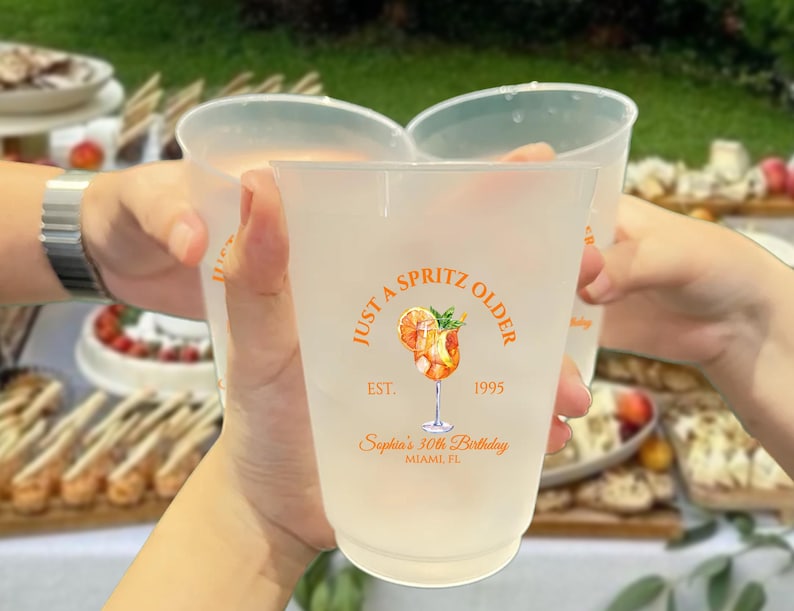 May include: Frosted plastic cups with orange text and a cocktail illustration are held up in a toast. The text on the cups reads "Just a Spritz Older" and "Sophia's 30th Birthday, Miami FL."