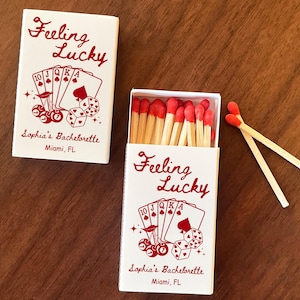 May include: Two white matchboxes with red text and graphics. One box is closed, the other open, revealing red-tipped matches. The text reads "Feeling Lucky" and "Sophia's Bachelorette, Miami, FL". The design includes playing cards and dice.
