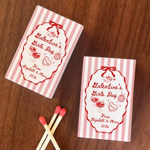 May include: Two matchboxes with pink and white stripes and red text that reads "Galentine's Girls Day" and "From Elizabeth & Olivia 2026". Three matches with red tips are also visible.