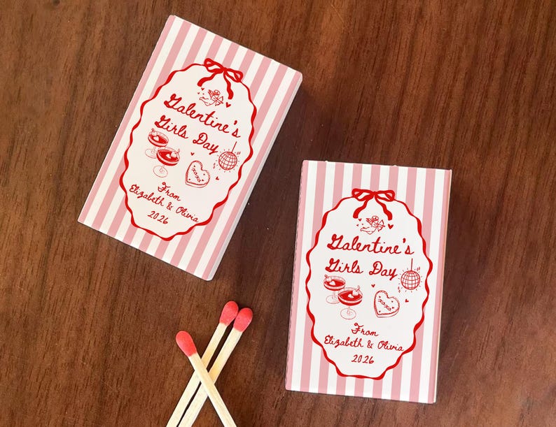 May include: Two matchboxes with pink and white stripes and red text that reads "Galentine's Girls Day" and "From Elizabeth & Olivia 2026". Three matches with red tips are also visible.