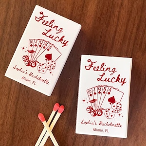 May include: Two white matchboxes with red text that reads "Feeling Lucky" and "Sophia's Bachelorette, Miami, FL." The boxes feature a poker-themed design with playing cards, dice, and cherries. Three matches with red tips are also visible.