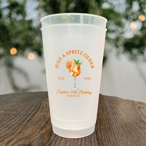 May include: A frosted plastic cup with the text "JUST A SPRITZ OLDER" in orange, along with a cocktail illustration. The cup also includes the text "EST. 1995" and "Sophia's 30th Birthday, Miami, FL".
