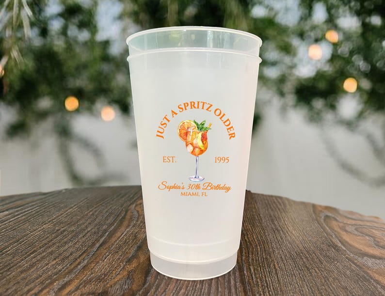 May include: A frosted plastic cup with the text "JUST A SPRITZ OLDER" in orange, along with a cocktail illustration. The cup also includes the text "EST. 1995" and "Sophia's 30th Birthday, Miami, FL".