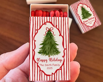 May include: A matchbox with a red and white striped design. The front features a green Christmas tree illustration inside a red decorative frame. The text "Happy Holidays The Smith Family 2025" is printed below the tree. Red match heads are visible.