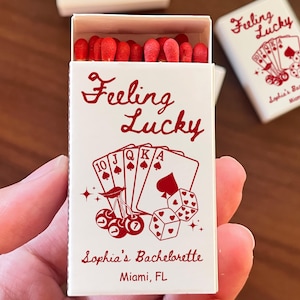 May include: A white matchbox with the words "Feeling Lucky" in red script, along with playing card illustrations and dice. The box also includes the text "Sophia's Bachelorette, Miami, FL". Red match heads are visible.