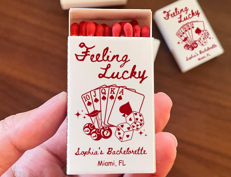 May include: A white matchbox with the words "Feeling Lucky" in red script, along with playing card illustrations and dice. The box also includes the text "Sophia's Bachelorette, Miami, FL". Red match heads are visible.