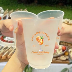 May include: Frosted plastic cups with the text "Just a Spritz Older" and a cocktail illustration in orange. The cups are being held up in a toast, with a blurred background of food and greenery. The text "Sophia's 30th Birthday, Miami FL" is also printed on the cups.