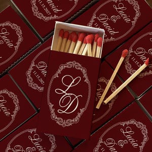 May include: A collection of rectangular matchboxes in deep red with white ornate oval frames. The boxes feature the names Lucia & David and the date 11.15.2027. One open box displays wooden matches with red tips, and two loose matches lie on top.