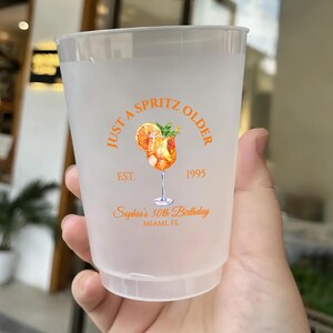May include: A frosted plastic cup with an orange and white design. The design includes the text "Just a Spritz Older" and "Sophia's 30th Birthday, Miami FL". The cup features a watercolor illustration of a cocktail.