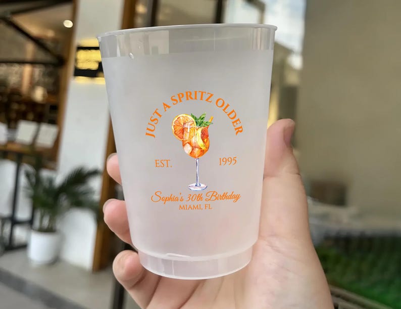 May include: A frosted plastic cup with an orange and white design. The design includes the text "Just a Spritz Older" and "Sophia's 30th Birthday, Miami FL". The cup features a watercolor illustration of a cocktail.
