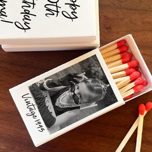 Op de afbeelding: Witte luciferdoos met een zwart-wit foto van een kind met een zonnebril en de tekst "Vintage 1995". De luciferkoppen zijn rood. De bovenkant van de doos heeft de tekst "Happy 30th Birthday, Anna!"