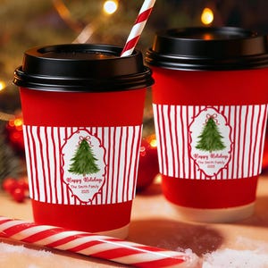 Può includere: Due tazze di caffè di carta rosse con coperchi neri, con una manica a righe bianche e rosse. Ogni manica presenta un disegno di albero di Natale con il testo "Happy Holidays The Smith Family 2025". Sono visibili anche un bastoncino di zucchero e una cannuccia a righe.