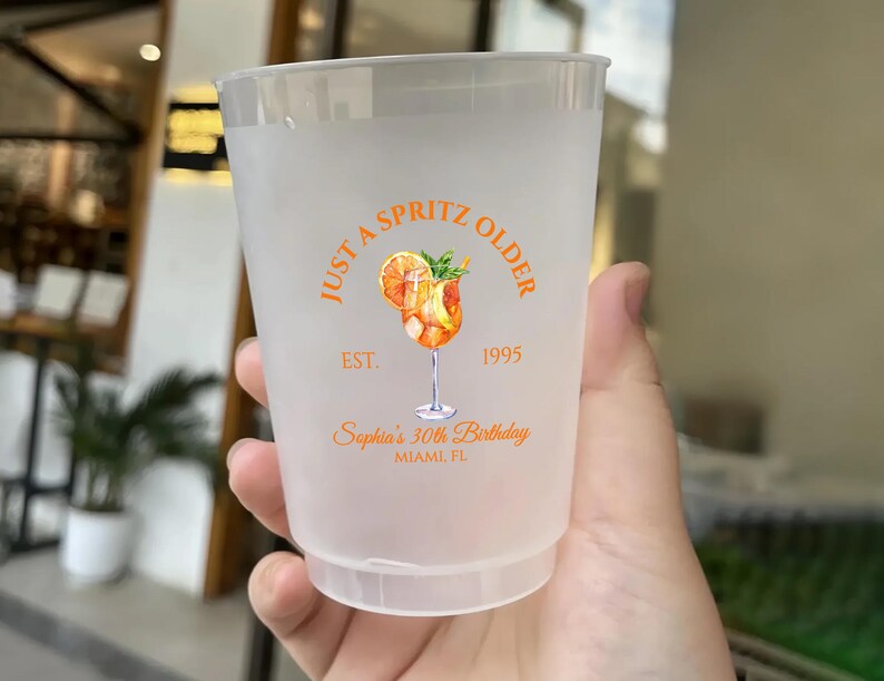 May include: A frosted plastic cup with an orange and white design. The design includes the text "Just a Spritz Older" and "Sophia's 30th Birthday, Miami FL". The cup features a watercolor illustration of a cocktail.