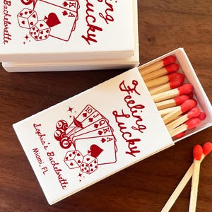 May include: White matchboxes with red text and illustrations, including playing cards and dice. One box is open, revealing matches with red tips. The text reads "Feeling Lucky" and "Sophia's Bachelorette, Miami, FL."