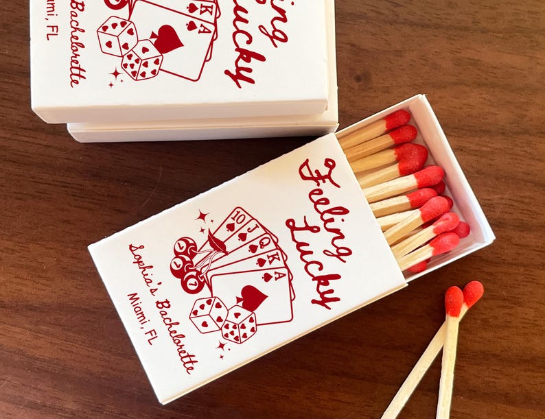 May include: White matchboxes with red text and illustrations, including playing cards and dice. One box is open, revealing matches with red tips. The text reads "Feeling Lucky" and "Sophia's Bachelorette, Miami, FL."