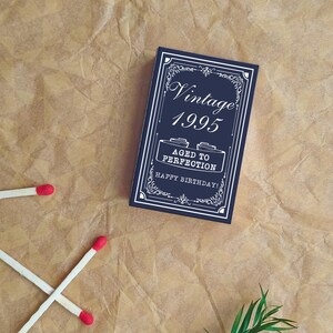 May include: A navy blue birthday card with white text that reads "Vintage 1995 Aged to Perfection Happy Birthday!" is on a brown paper background. Two matches are crossed, and another is nearby.