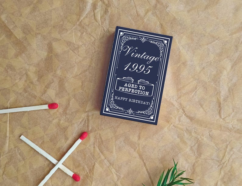 May include: A navy blue birthday card with white text that reads "Vintage 1995 Aged to Perfection Happy Birthday!" is on a brown paper background. Two matches are crossed, and another is nearby.