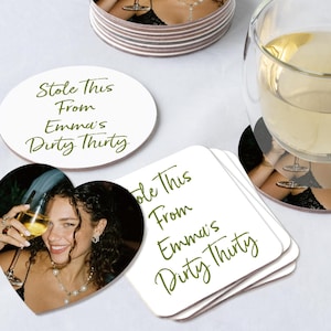 May include: White coasters featuring a photo of a woman with a glass of wine. The coasters have the text "Stole This From Emma's Dirty Thirty" in green script. One coaster is heart-shaped, another oval.