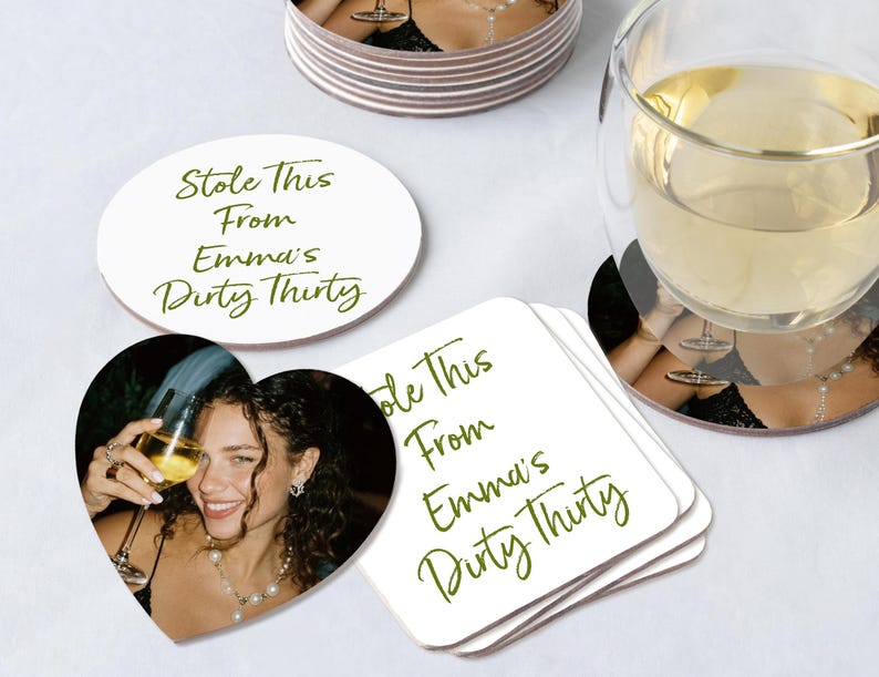 May include: White coasters featuring a photo of a woman with a glass of wine. The coasters have the text "Stole This From Emma's Dirty Thirty" in green script. One coaster is heart-shaped, another oval.