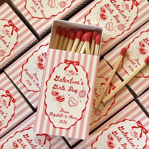 May include: A collection of pink and white striped matchboxes, each with a decorative label that reads "Valentine's Girls Day." The labels feature red illustrations and text, with an open box revealing red-tipped matches.