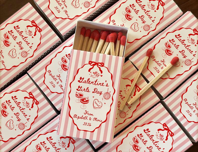 May include: A collection of pink and white striped matchboxes, each with a decorative label that reads "Valentine's Girls Day." The labels feature red illustrations and text, with an open box revealing red-tipped matches.
