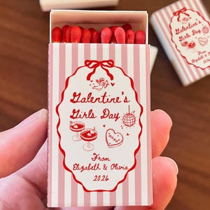 May include: A matchbox with a pink and white striped design, featuring the text "Galentine's Girls Day" in red script. The box is open, revealing red-tipped matches. The design includes illustrations of cocktails, a disco ball, and a heart-shaped cookie.