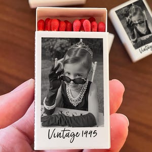 Op de afbeelding: Een witte luciferdoos met een zwart-wit foto van een kind met een zonnebril, tiara en handschoenen. De tekst "Vintage 1995" staat eronder. De luciferdoos bevat lucifers met rode koppen.