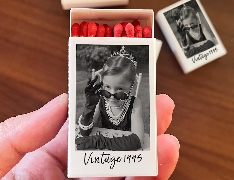 Op de afbeelding: Een witte luciferdoos met een zwart-wit foto van een kind met een zonnebril, tiara en handschoenen. De tekst "Vintage 1995" staat eronder. De luciferdoos bevat lucifers met rode koppen.