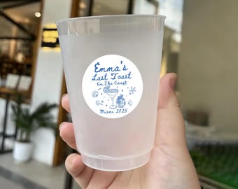 May include: A clear plastic cup with a white circular sticker featuring blue text and illustrations. The sticker reads "Emma's Last Toast On The Coast" with a cocktail, seashells, and the text "Miami 2025".