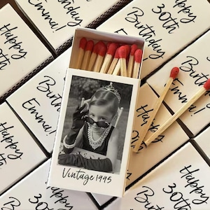 Op de afbeelding: Een verzameling witte lucifersdoosjes met zwarte tekst die luidt "Happy 30th Birthday Emma!". E&eacute;n doosje is open en toont lucifers met rode uiteinden en een zwart-witfoto van een kind met een zonnebril. De foto is gelabeld "Vintage 1995".