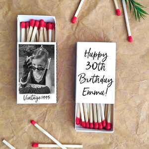 Op de afbeelding: Twee witte lucifersdoosjes met rood gekleurde lucifers. E&eacute;n doosje heeft een zwart-wit foto van een kind met een zonnebril en de tekst "Vintage 1995". Het andere doosje zegt "Happy 30th Birthday Emma!"