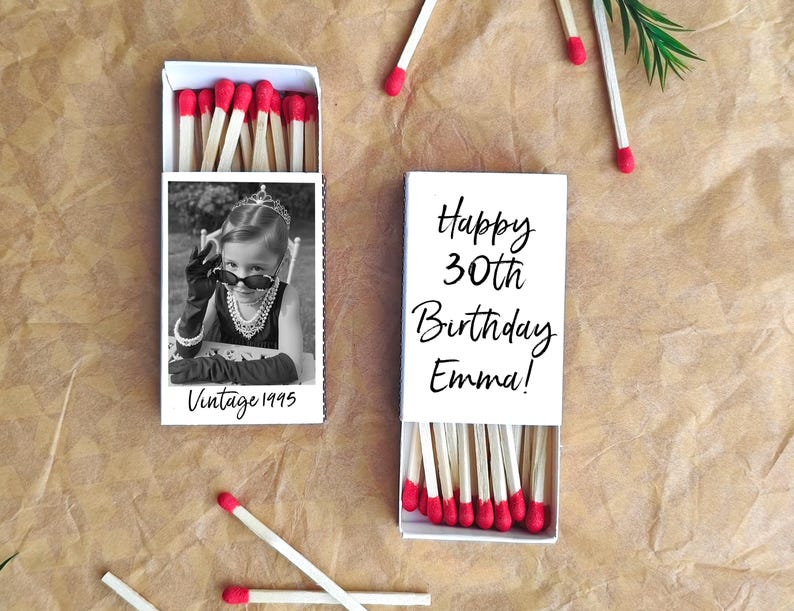 Op de afbeelding: Twee witte lucifersdoosjes met rood gekleurde lucifers. E&eacute;n doosje heeft een zwart-wit foto van een kind met een zonnebril en de tekst "Vintage 1995". Het andere doosje zegt "Happy 30th Birthday Emma!"