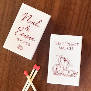 May include: Two white matchboxes with burgundy text and a few matches. One box reads "Noah & Emma" with a date and rings. The other box says "THE PERFECT MATCH" with swan illustrations.