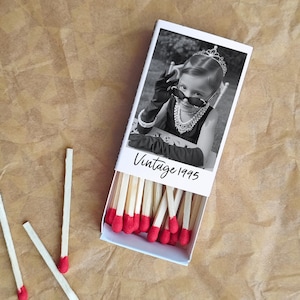Op de afbeelding: Een witte luciferdoos met een zwart-wit foto van een kind met een zonnebril en een tiara. De tekst "Vintage 1995" staat eronder. De doos is open en toont lucifers met rode uiteinden.