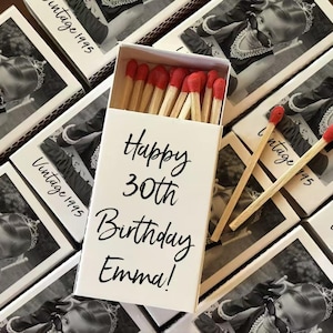 Op de afbeelding: Een witte luciferdoos met de tekst "Happy 30th Birthday Emma!" en meerdere dozen met een zwart-wit foto en de tekst "Vintage 1995". De luciferkoppen zijn rood.