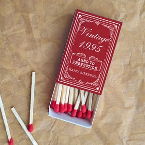 May include: A red matchbox with the words "Vintage 1995" and "Aged to Perfection Happy Birthday!" in white. The matchbox is open, revealing red-tipped matches. Three loose matches are scattered nearby. The background is a textured brown surface.
