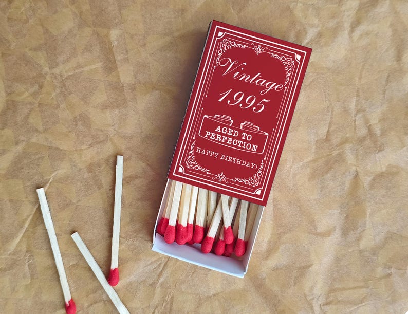 May include: A red matchbox with the words "Vintage 1995" and "Aged to Perfection Happy Birthday!" in white. The matchbox is open, revealing red-tipped matches. Three loose matches are scattered nearby. The background is a textured brown surface.