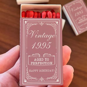 May include: A matchbox with a mauve cover, featuring the text "Vintage 1995" and "Aged to Perfection, Happy Birthday!" in white. The box is open, revealing red match heads. A second, closed box is visible.