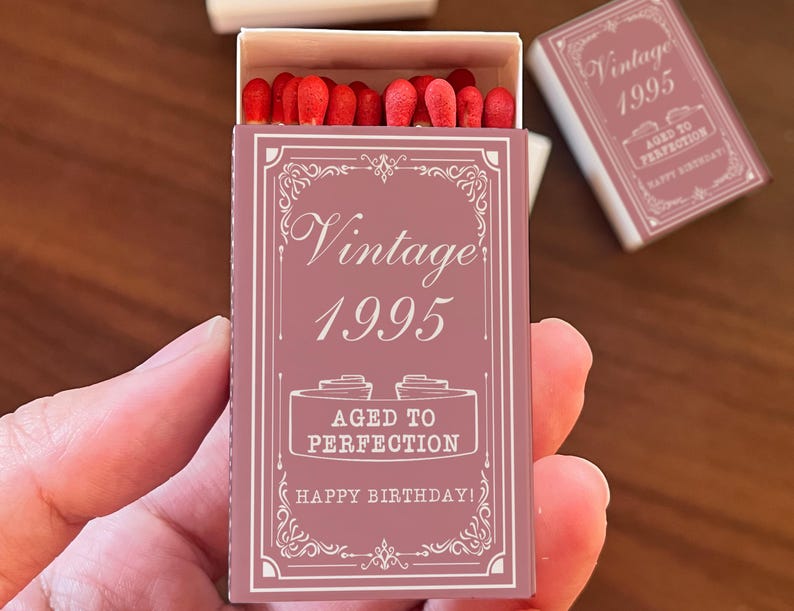 May include: A matchbox with a mauve cover, featuring the text "Vintage 1995" and "Aged to Perfection, Happy Birthday!" in white. The box is open, revealing red match heads. A second, closed box is visible.