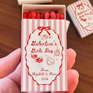May include: A matchbox with a pink and white striped design, featuring the text "Galentine's Girls Day" in red script. The box is open, revealing red-tipped matches. The design includes illustrations of cocktails, a disco ball, and a heart-shaped cookie.