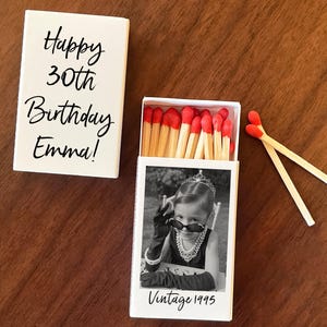 May include: A white matchbox with the text "Happy 30th Birthday Emma!" next to an open matchbox with a black and white photo of a girl wearing sunglasses and the text "Vintage 1995". Two matches are next to the open box.