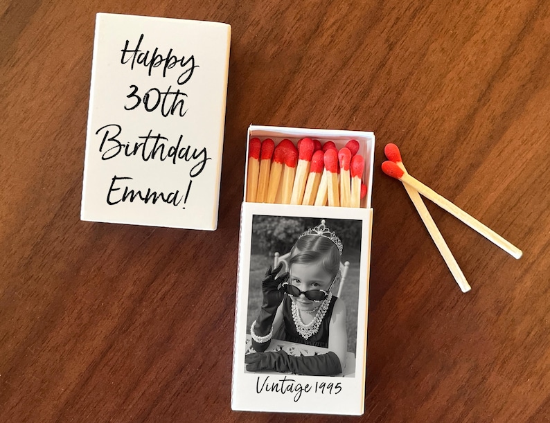 Op de afbeelding: Een witte luciferdoos met de tekst "Happy 30th Birthday Emma!" naast een open luciferdoos met een zwart-wit foto van een meisje met een zonnebril en de tekst "Vintage 1995". Twee lucifers liggen naast de open doos.